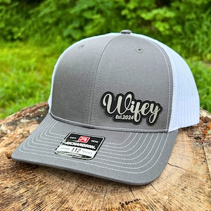 以下が含まれることがあります： グレーとホワイトのトラッカーハット。黒いパッチには「Wife」と「Est. 2024」の文字が書かれています。ハットにはリチャードソン112の調節可能なバックルが付いています。