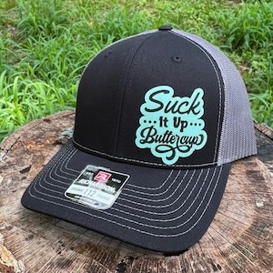 May include: Black and grey trucker hat with a turquoise blue patch that says "Suck It Up Buttercup" in black lettering.
