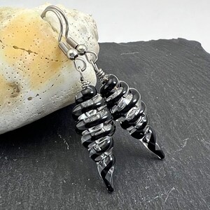May include: A pair of spiral-shaped earrings with alternating black and clear glass segments. The earrings have silver-colored ear wires and are displayed against a neutral background. The design is elegant and eye-catching, suitable for various occasions.