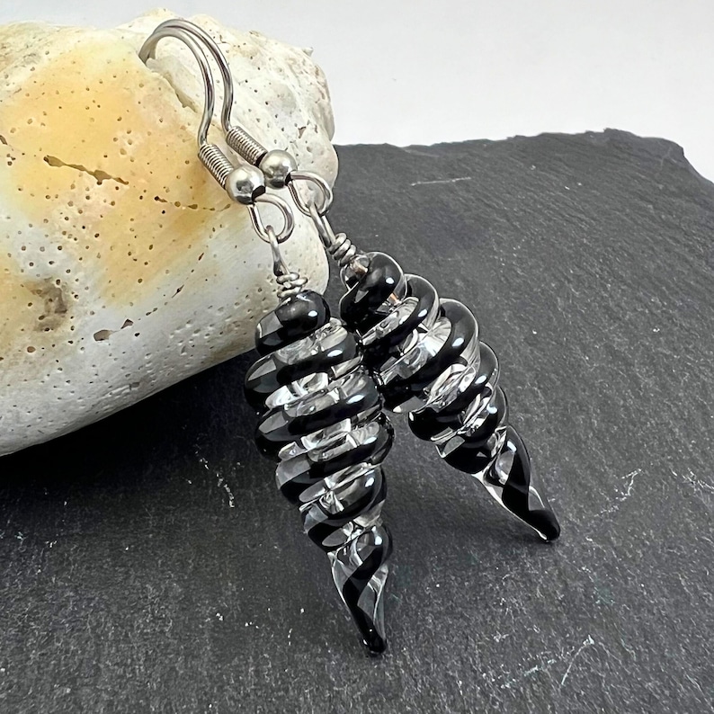 May include: A pair of spiral-shaped earrings with alternating black and clear glass segments. The earrings have silver-colored ear wires and are displayed against a neutral background. The design is elegant and eye-catching, suitable for various occasions.