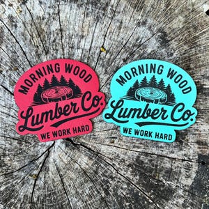 Puede incluir: Dos pegatinas con el texto "Morning Wood Lumber Co." y "We Work Hard". Una pegatina es roja, la otra es turquesa. Ambas tienen un gráfico negro de árboles y una rodaja de madera.
