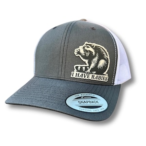 Op de afbeelding: Een grijze en witte truckercap met een zwart-witte wasbeer-afbeelding en de tekst "I Have Rabies". De pet heeft een wit mesh-achterpaneel en een grijs voorpaneel met een zwarte gestikte klep. De pet heeft een witte sticker aan de onderkant van de klep met de tekst "The Authentic Yupoong Snapback Yupoong Since 1974".