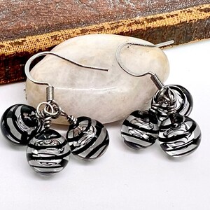 May include: A pair of dangle earrings featuring three black and silver striped glass beads. The beads are suspended from silver-colored ear wires. The earrings are displayed on a white, oval-shaped stone.