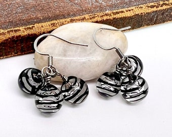May include: Dangle earrings with three black and silver striped glass beads. The beads are suspended from silver-coloured ear wires. The earrings are displayed on a white, oval-shaped stone.
