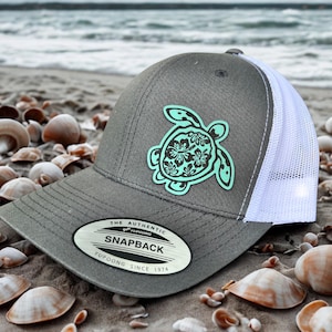Pode incluir: Boné snapback cinzento com costas em rede branca e um design de tartaruga marinha turquesa. O boné apresenta o texto "SNAPBACK" e "YUPOONG SINCE 1974" numa etiqueta. Numa praia arenosa com conchas.