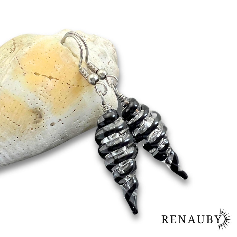May include: A pair of spiral-shaped earrings with black and clear glass, suspended from silver-colored hooks. The earrings are displayed against a textured, off-white surface. The brand name, RENAUBY, is visible in the lower right corner.