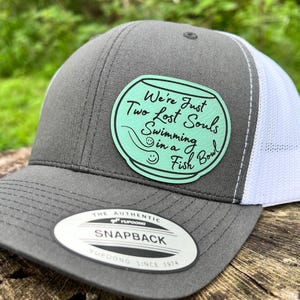 Könnte beinhalten: Graue Snapback-Kappe mit weißem Mesh-Rücken. Auf der Kappe befindet sich ein kreisförmiger Aufnäher mit dem Text "We're Just Two Lost Souls Swimming in a Fish Bowl". Der Schirm hat eine gebogene Form und einen Snapback-Verschluss.