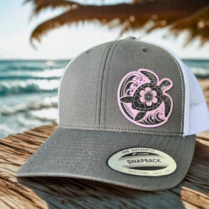 May include: A grey snapback cap with a white mesh back. The cap features a pink and black turtle design with a flower on the front. The cap has a curved peak and a snap closure. The tag reads "The Authentic Snapback Yupoong Since 1974."