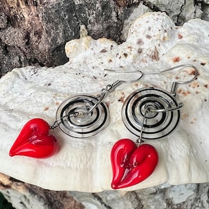 May include: A pair of dangle earrings featuring a spiral design with black and clear accents. Each earring has a vibrant red heart-shaped charm. The earrings are suspended from silver-colored ear wires.