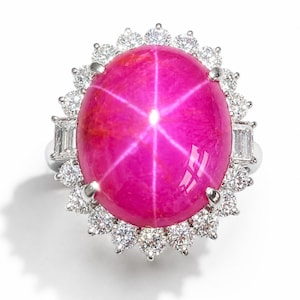 May include: A ring featuring a large, oval, cabochon-cut hot pink star ruby as the centerpiece. The ruby displays a distinct six-rayed star effect. It is surrounded by a halo of round brilliant-cut diamonds and two baguette-cut diamonds.