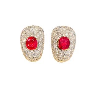 May include: A pair of oval-shaped earrings. Each earring features a vibrant red gemstone at the center, surrounded by a halo of smaller, sparkling stones. The setting appears to be gold-toned, creating a luxurious look.