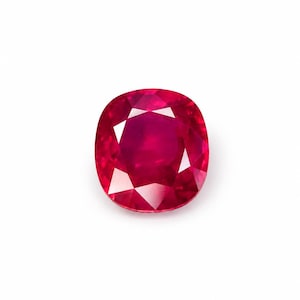 May include: A cushion-cut ruby gemstone with a deep, saturated red color and multiple triangular and trapezoidal facets that create a reflective, brilliant surface. The stone has a rounded square shape and a transparent, polished appearance.