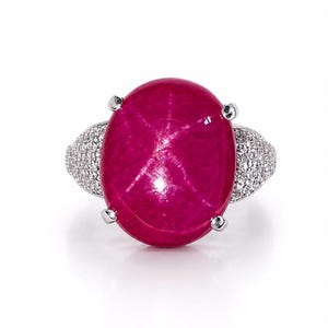 Op de afbeelding: Een ring met een grote, ovaalvormige, dieproze sterrobijn cabochon in een zilverkleurige metalen zetting met pootjes. De ringband is bezet met talloze kleine, heldere, fonkelende steentjes die langs de zijkanten van de ring doorlopen.