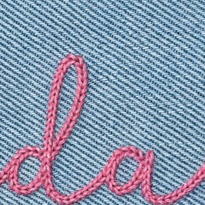 May include: Close-up of denim fabric with pink embroidered cursive lettering. The pink thread is stitched in a chain-like pattern, creating a textured effect against the blue denim background. The lettering appears to be part of a word.