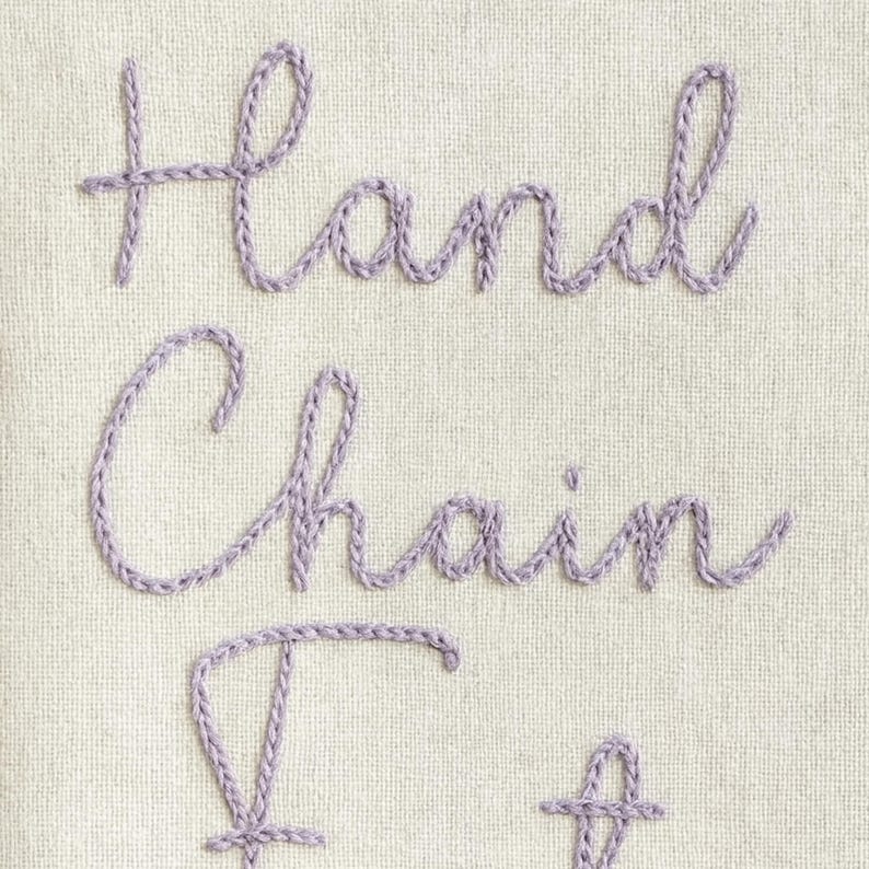 May include: Close-up of a cream-colored fabric with the embroidered words "Hand Chain". The text is in a lavender color, with a cursive font. The embroidery thread has a textured, rope-like appearance.