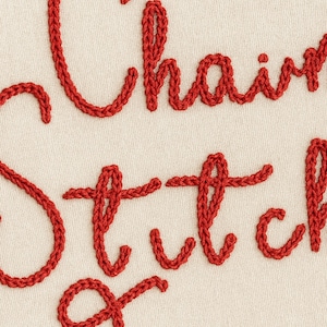 Peut inclure: Gros plan sur un tissu avec les mots "Chain Stitch" brodés en fil rouge. Les lettres sont dans un style cursif, et le fond est de couleur crème. La couture est épaisse et texturée.