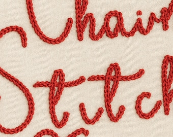 May include: Close-up of fabric with the words "Chain Stitch" embroidered in red thread. The letters are in a cursive style, and the background is a cream color. The stitching is thick and textured.