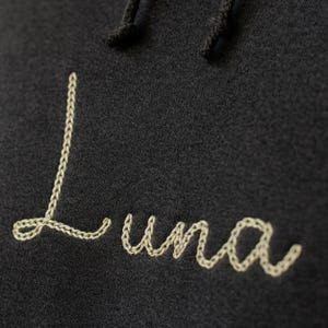 Puede incluir: Primer plano de una sudadera con capucha gris oscuro con cord&oacute;n. La palabra "Luna" est&aacute; bordada en cursiva con hilo beige claro. El bordado tiene apariencia de punto de cadeneta.