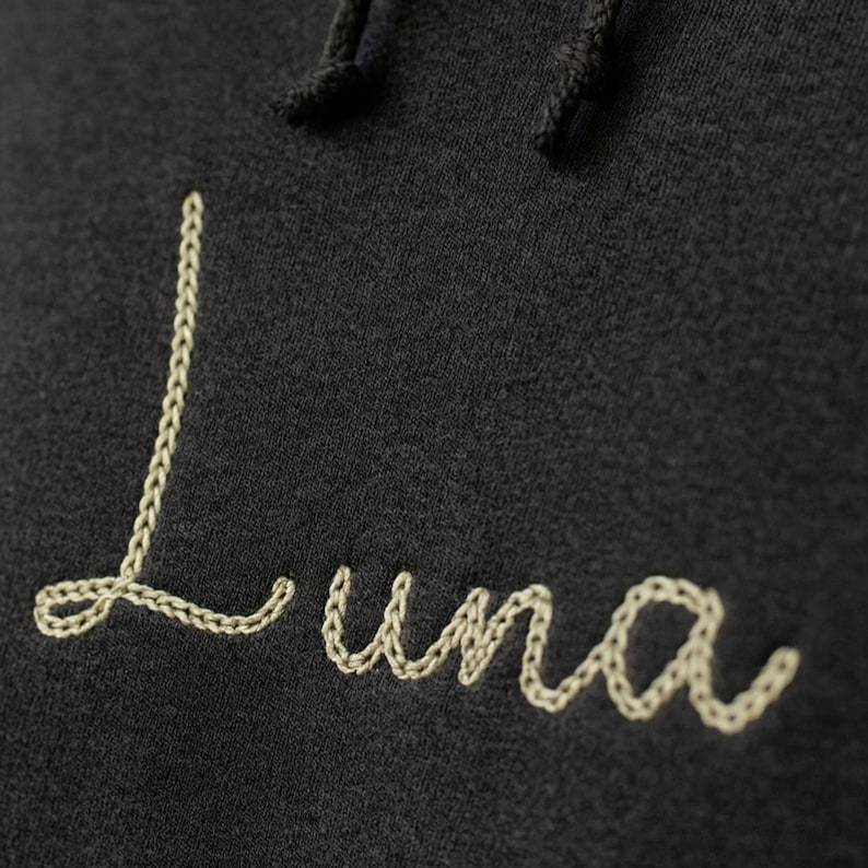 Puede incluir: Primer plano de una sudadera con capucha gris oscuro con cord&oacute;n. La palabra "Luna" est&aacute; bordada en cursiva con hilo beige claro. El bordado tiene apariencia de punto de cadeneta.