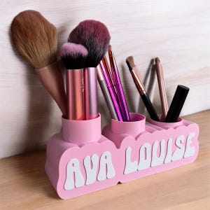 May include: Pink makeup brush holder with three compartments. The holder has the names "Ava Louise" written in white on the front.