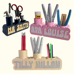 May include: Three personalised desk organisers with different coloured bases and names. The first is navy blue with the name "MR SMITH" in yellow letters. The second is pink with the name "AVA LOUISE" in white letters. The third is beige with the name "TILLY WILLOW" in silver letters.
