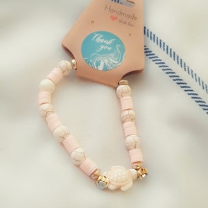 May include: A pink and white beaded bracelet with a gold turtle charm. The bracelet is handmade and comes with a tag that says "Handmade with love" and "Thank you".