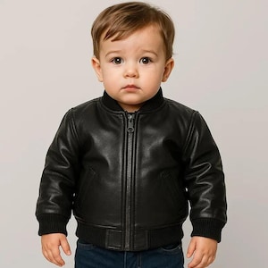 May include: A black leather bomber jacket for a toddler. The jacket features a ribbed collar, cuffs, and hem, a front zipper closure, and two front pockets. The child is wearing the jacket and looking at the camera.