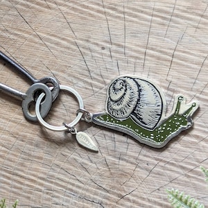May include: A silver key ring with two antique keys and a green snail charm. The charm is detailed with a black and white snail shell and a green body. The keyring is on a wooden surface with green ferns.