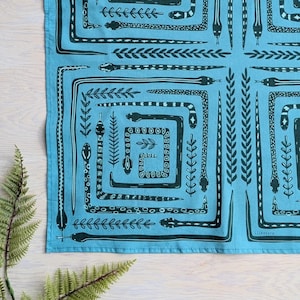 May include: A square, turquoise textile with a repeating pattern of dark teal snakes and botanical elements. The design features snakes in a square formation, with various leaf and plant motifs. The textile is laid flat on a light wood surface.