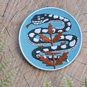 May include: A circular patch with a black and white snake design on a light blue background. The snake is coiled around a brown plant. The patch is set on a wooden surface with green fern leaves.