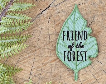 Può includere: Una toppa verde a forma di foglia con il testo "FRIEND of the FOREST" ricamato in filo nero. La toppa è su una superficie di legno con una felce.