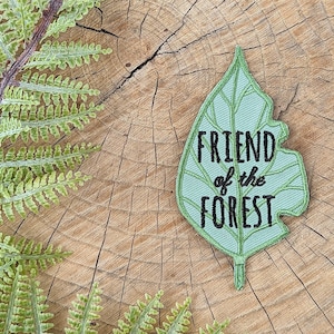 May include: A green leaf-shaped patch with the text "FRIEND of the FOREST" embroidered in black thread. The patch is on a wooden surface with a fern.