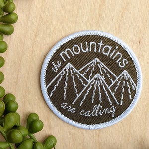 May include: Round embroidered patch with the text "the mountains are calling" and a mountain graphic. The patch has a brown background, a light blue border, and white stitching. Green plant sprigs are in the background.