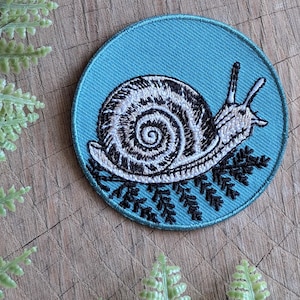 May include: A blue embroidered patch with a white snail on a bed of green leaves. The snail has a black shell with a white spiral.