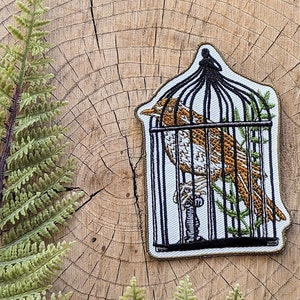 May include: A brown and white bird is trapped inside a black wire cage with a white background. The cage is embroidered on a white fabric patch with a gold outline.