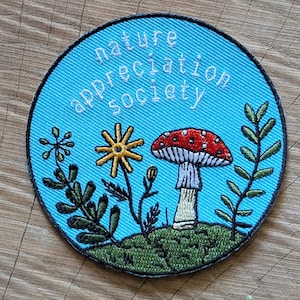 May include: Embroidered patch with a light blue background featuring the text "nature appreciation society". The patch includes a red and white mushroom, a yellow flower, and green foliage. The patch is circular in shape.