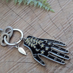 May include: A silver keychain with a black hand charm featuring gold stars, moons, and leaves. The charm is attached to a silver ring and a silver tag that reads "likesara".