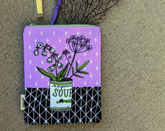 May include: A purple zippered pouch with a pink background and a black and white geometric pattern. The pouch has a design of a can of soup with flowers and the words "organic soup condensed" on it.