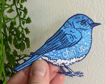 Può includere: Toppa ricamata blu a forma di uccello con il testo "chin up" sul petto.