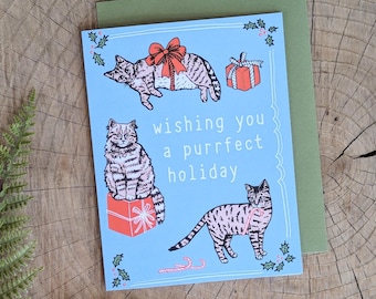 May include: A blue Christmas card with a festive message that reads "Wishing you a purrfect holiday". The card features three illustrations of cats in various poses, including one cat sitting in a gift box, one cat lying down, and one cat standing up. The card has a green border and a red bow.