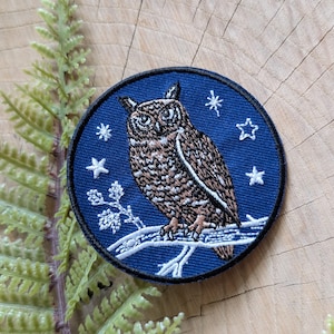 May include: A circular embroidered patch featuring a brown owl perched on a white branch against a dark blue background. The design includes white stitched stars and small floral accents surrounding the owl, finished with a black stitched border.
