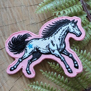 May include: A pink embroidered patch featuring a running horse with a black mane and tail. The horse is white with black detailing and a blue star on its flank. The patch is on a wooden surface with green fern.