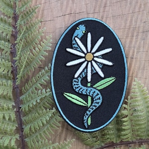 May include: An embroidered patch featuring a blue and white snake entwined around a white daisy with a yellow center. The design is set against a black oval with a light blue border. Green leaves are also included in the design.