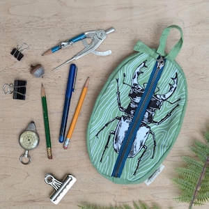 May include: A green pencil case with a zipper closure and a graphic print of a stag beetle. The pencil case is on a wooden surface with various school supplies.