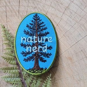 Puede incluir: Un parche bordado de forma ovalada con un fondo azul brillante y un diseño de pino en color marrón oscuro y negro. Las palabras "nature nerd" están bordadas con hilo blanco sobre el centro del árbol. El parche tiene un borde verde.