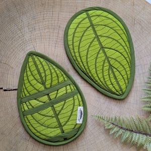 May include: Two leaf-shaped, quilted pot holders in shades of green. The pot holders have a leaf vein pattern and a darker green trim. One pot holder has two additional green straps. The pot holders are on a wooden surface.