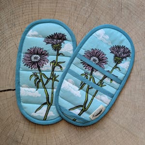 May include: Two quilted oven mitts with a light blue background featuring a floral design with purple flowers and white clouds. The mitts have a teal blue trim and a quilted pattern. The design includes green stems and leaves.