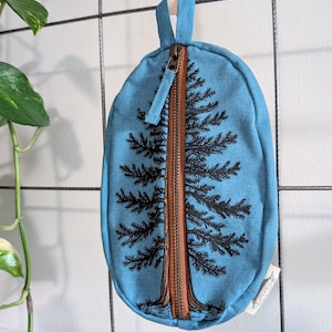 Pu&ograve; includere: Una custodia ovale in tela blu con una cerniera marrone verticale centrale che imita il tronco di un pino nero illustrato. I rami dell'albero si estendono sul tessuto blu su entrambi i lati della cerniera. &Egrave; presente una piccola etichetta bianca.