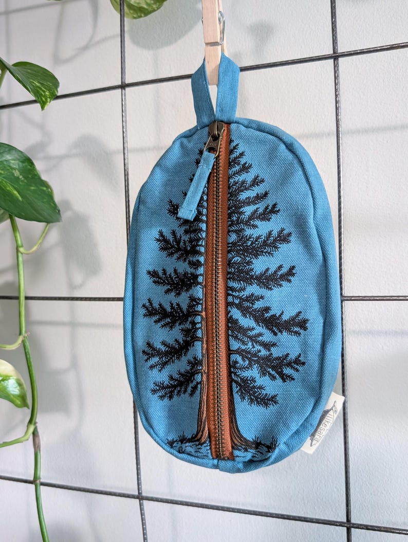 Pu&ograve; includere: Una custodia ovale in tela blu con una cerniera marrone verticale centrale che imita il tronco di un pino nero illustrato. I rami dell'albero si estendono sul tessuto blu su entrambi i lati della cerniera. &Egrave; presente una piccola etichetta bianca.