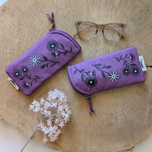 May include: Two purple eyeglass cases with a floral design and a pair of clear-framed glasses on a wooden surface. The cases have a zipper closure and a black and white floral pattern. The glasses are a light brown colour.